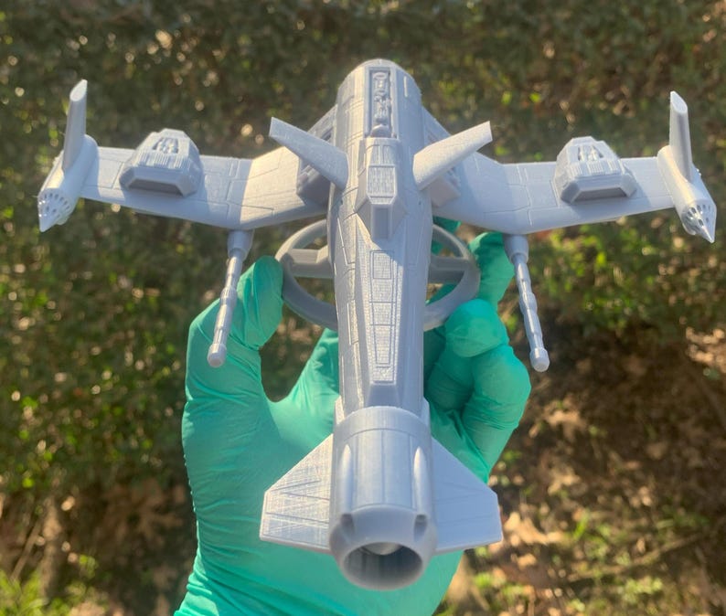 May include: A gray 3D printed model of a futuristic spaceship with a long, cylindrical body and two wings extending out to the sides. The spaceship has a detailed design with a variety of panels and hatches.