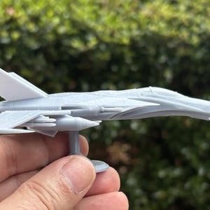 Puede incluir: Un modelo gris de avi&oacute;n, probablemente un avi&oacute;n de combate, se sostiene en una mano. El avi&oacute;n en miniatura presenta alas, aletas de cola y un soporte detallados. El modelo est&aacute; hecho de un material gris claro sobre un fondo verde borroso.