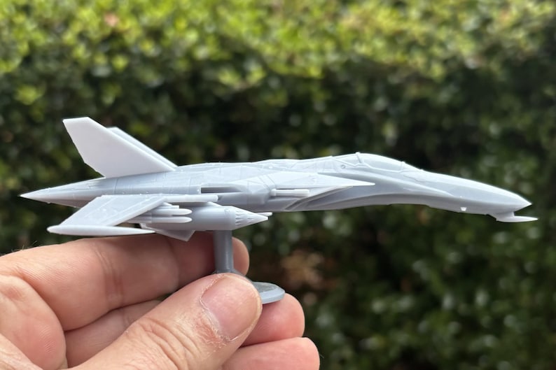 Puede incluir: Un modelo gris de avi&oacute;n, probablemente un avi&oacute;n de combate, se sostiene en una mano. El avi&oacute;n en miniatura presenta alas, aletas de cola y un soporte detallados. El modelo est&aacute; hecho de un material gris claro sobre un fondo verde borroso.
