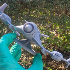 May include: A gray 3D printed model of a spaceship with a large engine in the back and two wings. The model is sitting on a stand and is being held by a gloved hand.