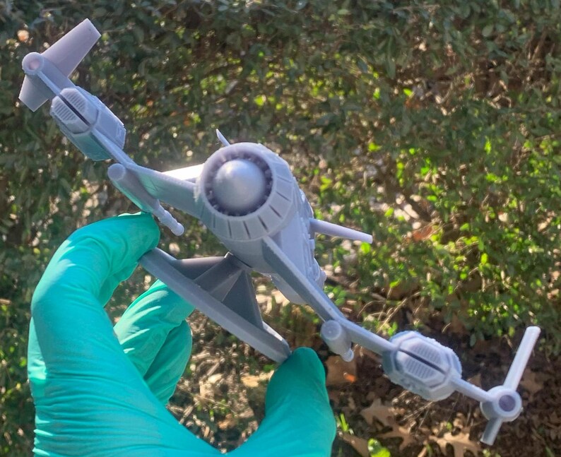 May include: A gray 3D printed model of a spaceship with a large engine in the back and two wings. The model is sitting on a stand and is being held by a gloved hand.