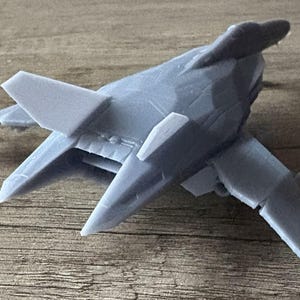 Puede incluir: Un modelo de avi&oacute;n gris con un dise&ntilde;o &uacute;nico, con alas anguladas y una distintiva aleta de cola. El avi&oacute;n en miniatura est&aacute; hecho de un material s&oacute;lido y se encuentra sobre una superficie de madera. El modelo es una representaci&oacute;n detallada de una aeronave futurista.