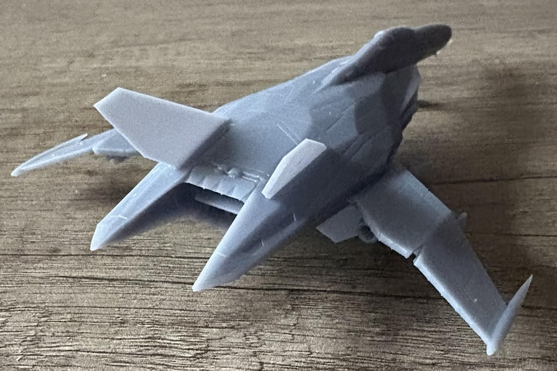 Puede incluir: Un modelo de avi&oacute;n gris con un dise&ntilde;o &uacute;nico, con alas anguladas y una distintiva aleta de cola. El avi&oacute;n en miniatura est&aacute; hecho de un material s&oacute;lido y se encuentra sobre una superficie de madera. El modelo es una representaci&oacute;n detallada de una aeronave futurista.