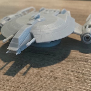 Può includere: Un modello di nave spaziale stampato in 3D di colore grigio con una parte superiore arrotondata a forma di disco e un corpo lungo e stretto. La nave spaziale ha un design dettagliato con più alette e un grande motore sul retro.