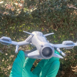 May include: A gray 3D printed model of a futuristic spaceship with a single large engine in the front and two smaller engines on the wings. The spaceship has a detailed design with a cockpit and a variety of other features.