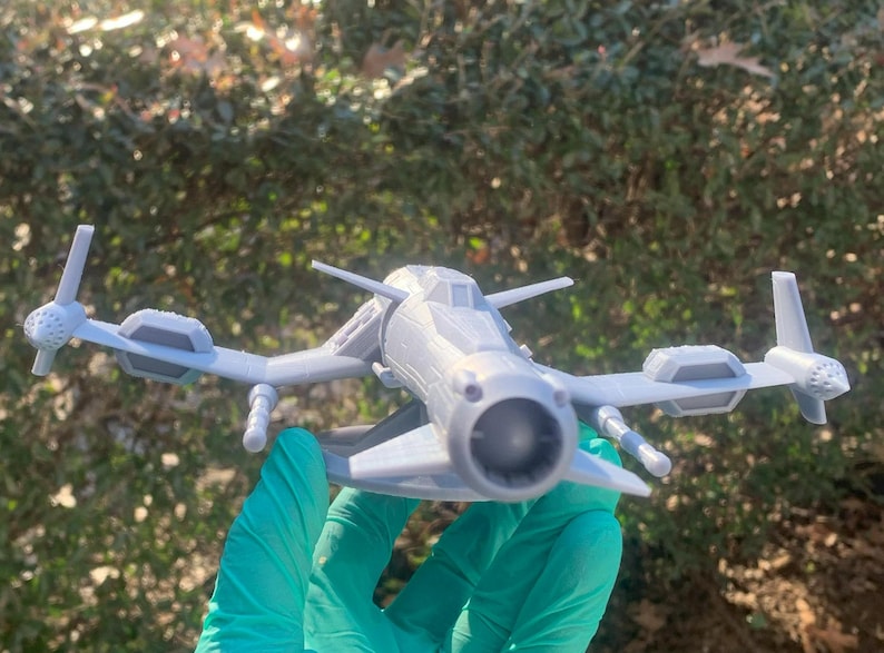 May include: A gray 3D printed model of a futuristic spaceship with a single large engine in the front and two smaller engines on the wings. The spaceship has a detailed design with a cockpit and a variety of other features.
