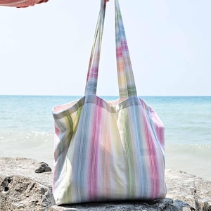 Puede incluir: Un bolso tote a rayas en tonos pastel de rosa, verde, amarillo y azul. El bolso se sostiene por sus largas correas, mostrando su diseño espacioso. El fondo presenta el mar y el cielo.
