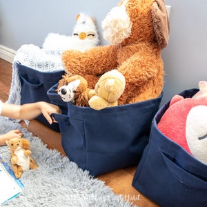 Puede incluir: Tres cestas de almacenamiento de lona azul marino llenas de animales de peluche. Las cestas tienen un bolsillo en el lateral. Una mano de niño alcanza una de las cestas.