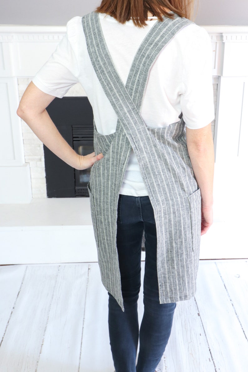 May include: A grey and white pinstriped apron featuring a cross-back design with wide shoulder straps and two side pockets. The apron is worn over a white short-sleeved shirt and dark blue jeans, showcasing the back view of the garment.
