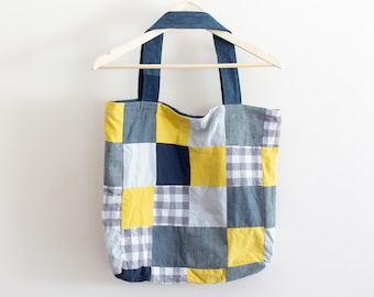 May include: A patchwork tote bag with blue denim, yellow, and gray checkered fabric. The bag has two denim straps and is hanging on a wooden hanger.