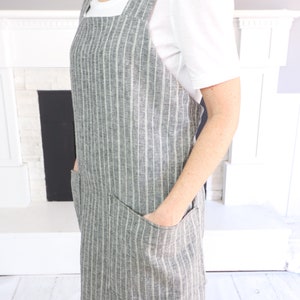May include: A gray and white pinstriped apron featuring a square neckline, wide shoulder straps, and two large patch pockets on the front. The apron is constructed with a textured fabric and has a straight hemline that falls to the mid-thigh area.