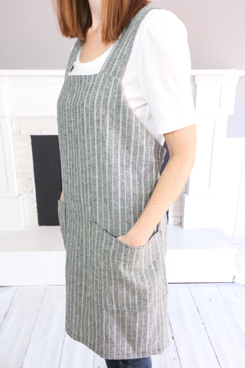May include: A gray and white pinstriped apron featuring a square neckline, wide shoulder straps, and two large patch pockets on the front. The apron is constructed with a textured fabric and has a straight hemline that falls to the mid-thigh area.