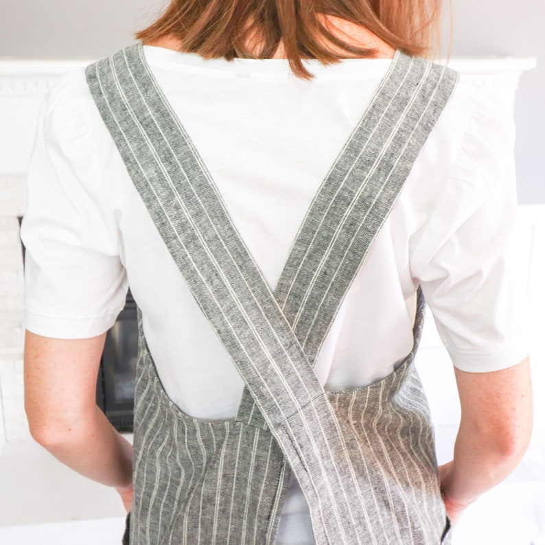 May include: A white short-sleeved shirt worn under a grey and white striped apron with wide shoulder straps that cross over the back. The apron features a textured fabric with vertical pinstripes and a simple design that covers the torso area.