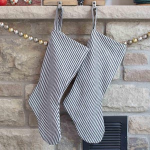 Könnte beinhalten: Zwei Weihnachtsstrümpfe mit einem schwarz-weiß gestreiften Muster hängen an Metallhaken an einem Steinkamin. Die Strümpfe sind aus Stoff und haben ein schlichtes, klassisches Design. Eine dekorative Girlande mit goldenen und silbernen Perlen hängt in der Nähe.