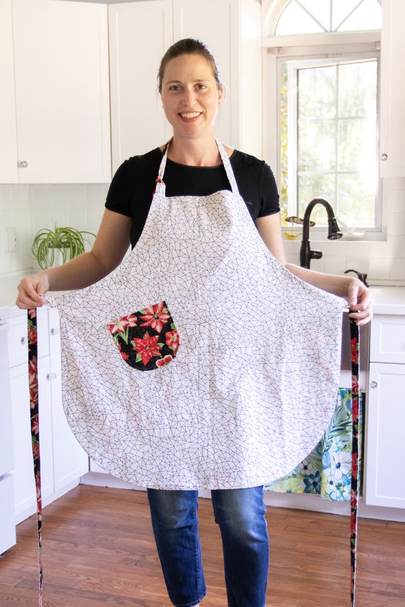 May include: A white apron with a black geometric pattern and a red and black floral pocket. The apron has long ties and is being held by a person.