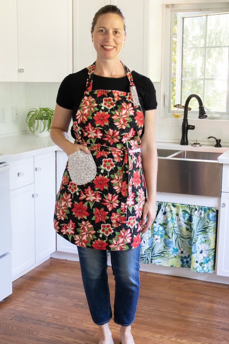 May include: A woman wearing a black apron with a red and green floral print. The apron has a white pocket with a geometric pattern. She is standing in a kitchen with a white countertop and a stainless steel sink.