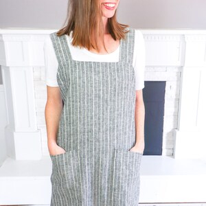 May include: A charcoal gray apron featuring a white vertical pinstripe pattern, a square neckline, wide shoulder straps, and two large rectangular front pockets. The apron is worn over a plain white short-sleeved t-shirt and dark blue denim jeans.
