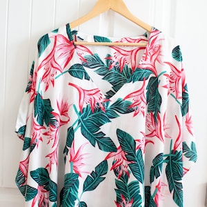 May include: White, loose-fitting, short-sleeved shirt with a V-neck and a tropical print featuring pink flowers and green leaves.