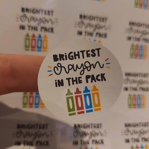 Könnte beinhalten: Ein runder Aufkleber mit dem Text "BRIGHTEST crayon IN THE PACK" in schwarzer Schrift. In der Mitte des Aufklebers sind vier Stifte gezeichnet, grün, rot, orange und blau.