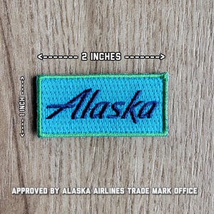 May include: A blue and green embroidered patch with the word "Alaska" in black cursive lettering. The patch measures 2 inches wide and 1 inch tall.