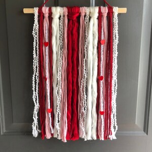 May include: A red, white, and pink yarn wall hanging with red heart accents. The yarn is woven together to create a textured and layered look.
