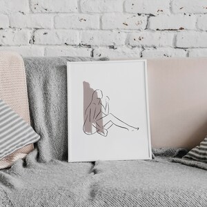 Könnte beinhalten: Ein gerahmter Line-Art-Druck einer sitzenden Person mit einem zarten rosafarbenen Hintergrund. Der Druck befindet sich auf einem grauen Sofa mit Kissen.
