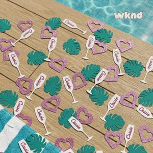 May include: Party decorations scattered on a wooden surface. The decorations include green tropical leaves, pink glitter heart shapes, and white champagne flutes with the name "Cassie" printed on them. A blue and white striped towel is in the lower left corner.