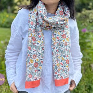 May include: A floral scarf with a white base, featuring red, yellow, and blue flowers. The scarf has a coral border and is worn around the neck. The word "mauve" is in a purple circle in the lower left corner of the image.