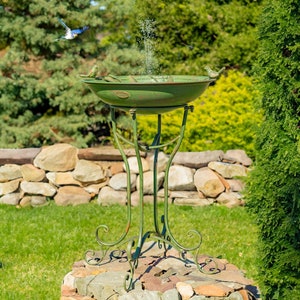May include: A green metal bird bath with a fountain and a decorative stand. The bird bath is on a stone base and surrounded by green grass.