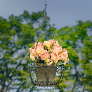 May include: A hanging planter with a metal frame and a rustic brown pot filled with a bouquet of pink artificial roses.