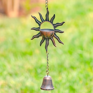 May include: A copper-toned metal wind chime featuring a sun design with a bell hanging from the bottom. The sun has a hollow center and is surrounded by rays.