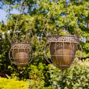 May include: Two hanging planters with a metallic bronze finish, featuring a wire cage design and an ornate, crown-like rim. Each planter is suspended by a metal chain and contains a solid, bowl-shaped inner container for holding plants.