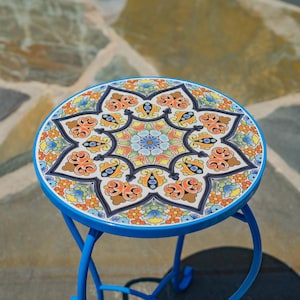 May include: A round side table with a blue metal frame and a circular top featuring a vibrant, multi-colored floral and geometric tile pattern in shades of orange, yellow, green, and blue, arranged in a symmetrical star-like design.