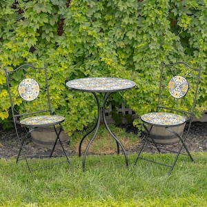 May include: A black metal bistro set with a round table and two folding chairs. The table and chair seats have a mosaic tile design with yellow, blue, and white floral patterns. The chairs have decorative backrests with matching tile accents. The set is on a grassy lawn.