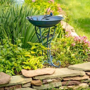 May include: A blue metal bird bath with a decorative bird perched on the rim. The bird bath has a round basin and sits on a curved, ornate stand. The bird bath is surrounded by green plants and flowers.