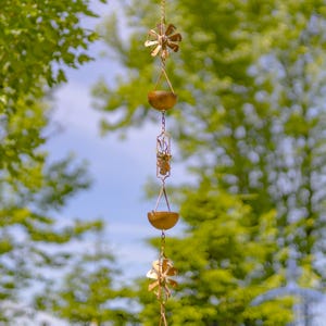 May include: A gold-colored rain chain with four decorative windmills and three bowls. The chain is hanging from a tree branch against a blurred background of green leaves.