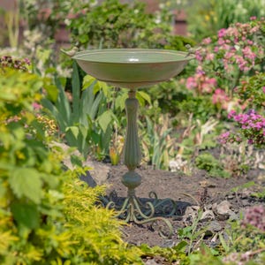 May include: A sage green birdbath featuring a wide, shallow basin with two small bird figurines perched on opposite rims. The basin is supported by a slender, fluted pedestal base that rests on a decorative, scrolled metal stand with four feet.