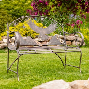 May include: Bronze-colored metal garden bench with an eagle and mountain design. The bench features a diamond-patterned back with stars and a detailed eagle in flight. The bench is set on a grassy lawn with a blurred background of flowers and foliage.