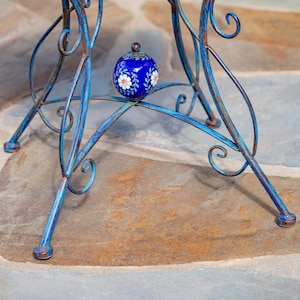 Puede incluir: Un peque&ntilde;o objeto decorativo redondo de color azul cobalto con dise&ntilde;os florales blancos y un peque&ntilde;o pomo marr&oacute;n descansa sobre una mesa de metal azul. La mesa tiene un acabado envejecido y un dise&ntilde;o en espiral. Las patas de la mesa son de un color azul similar.