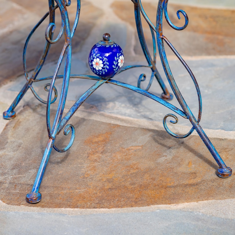 Puede incluir: Un peque&ntilde;o objeto decorativo redondo de color azul cobalto con dise&ntilde;os florales blancos y un peque&ntilde;o pomo marr&oacute;n descansa sobre una mesa de metal azul. La mesa tiene un acabado envejecido y un dise&ntilde;o en espiral. Las patas de la mesa son de un color azul similar.