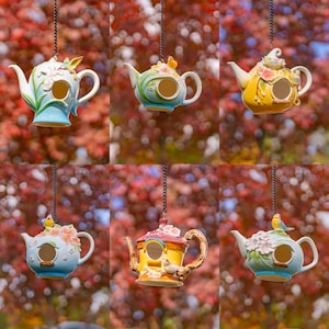 May include: A collection of six decorative ceramic birdhouses shaped like teapots. Each teapot birdhouse is painted in a different color scheme, including blue, yellow, and green, and features floral and bird accents. They hang from chains.