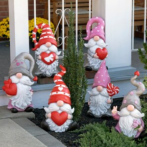 May include: Seven Valentine's Day gnomes with pink and red hats, holding hearts and candy canes. The gnomes are standing on a porch with a green bush behind them.