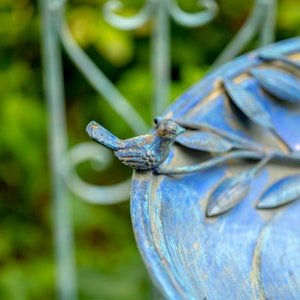 May include: A close-up of a blue bird bath with a decorative metal bird perched on the edge. The bird bath has a weathered, aged appearance with blue and bronze tones. The background is blurred, showing green foliage and a wrought iron fence.