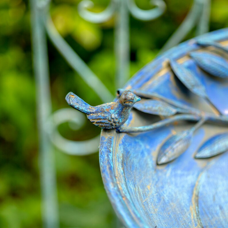 May include: A close-up of a blue bird bath with a decorative metal bird perched on the edge. The bird bath has a weathered, aged appearance with blue and bronze tones. The background is blurred, showing green foliage and a wrought iron fence.
