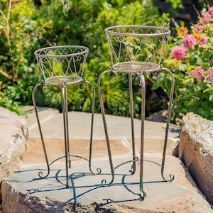 May include: Two metal plant stands with decorative scrollwork. The larger stand is taller and has a wider base. Both stands are made of a dark brown metal.