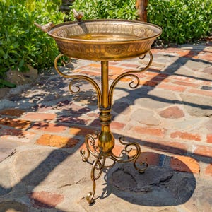 May include: A golden bird bath with a decorative, ornate design. The bird bath has a round basin with embossed detailing and a small bird perched on the rim. The stand features elegant, curved supports and a three-legged base.