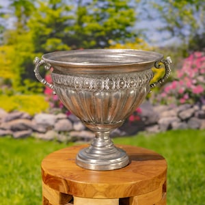 May include: A silver-toned metal urn with a wide, flared rim, a fluted body, and two ornate, curved handles on the sides. The upper section of the urn features a decorative band with a repeating circular pattern, supported by a pedestal base.