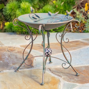 May include: A decorative bird bath with a weathered green finish and bronze accents. The metal bird bath features a shallow basin with leaf and bird details, supported by a scrolled metal stand. A decorative ceramic sphere is suspended from the stand.