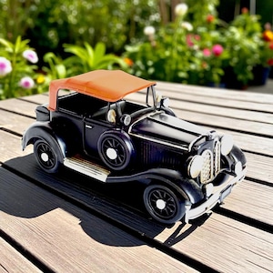 May include: A black vintage car model with a tan convertible top. The car has white-rimmed wheels and chrome accents. The model is sitting on a wooden surface, with a blurred background of greenery and flowers.