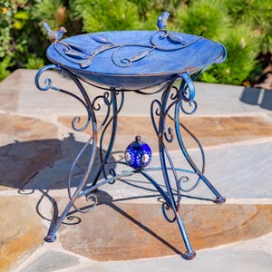 Puede incluir: Un bebedero para p&aacute;jaros de metal azul con un dise&ntilde;o decorativo. El bebedero tiene una cuenca redonda con detalles de hojas y p&aacute;jaros, sostenida por un soporte de metal en espiral. Una peque&ntilde;a esfera floral azul y blanca est&aacute; suspendida debajo.
